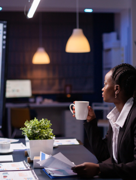 Workaholic african american businesswoman analysing marketing statistics on paperwork drinking coffee sitting at table late at night. Business manager with dark skin working in office meeting room