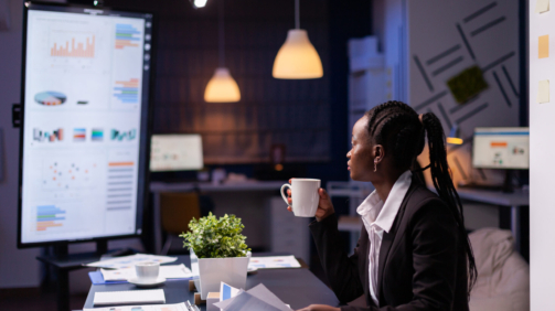 Workaholic african american businesswoman analysing marketing statistics on paperwork drinking coffee sitting at table late at night. Business manager with dark skin working in office meeting room